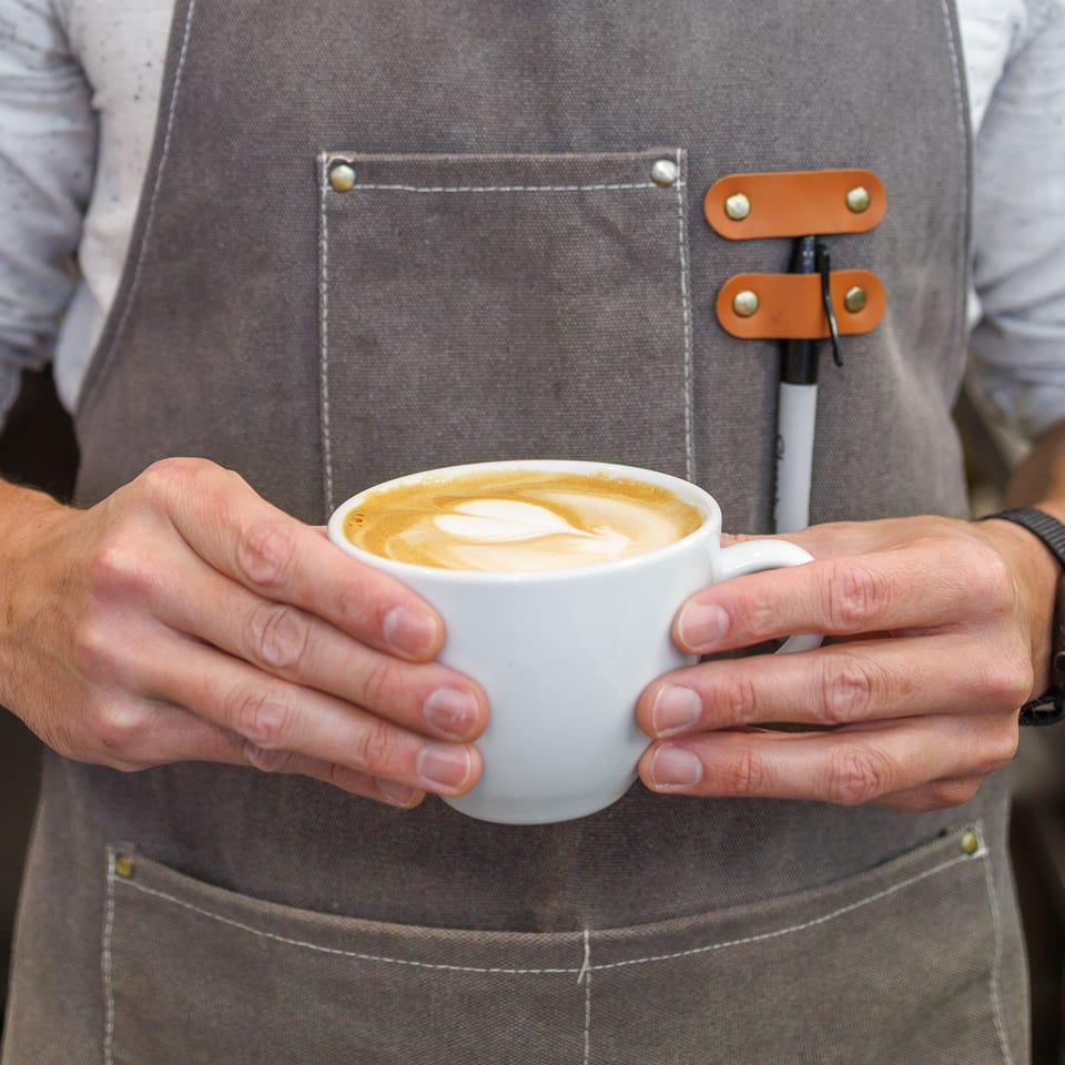 Barista in a gray apron holding a white cup of latte with heart-shaped art.