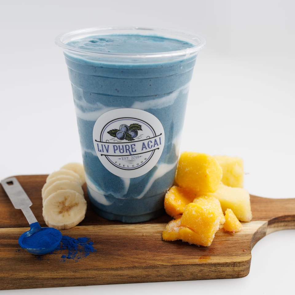 Blue layered açaí bowl in a branded cup with banana slices, mango chunks, and blue powder on a wooden board.