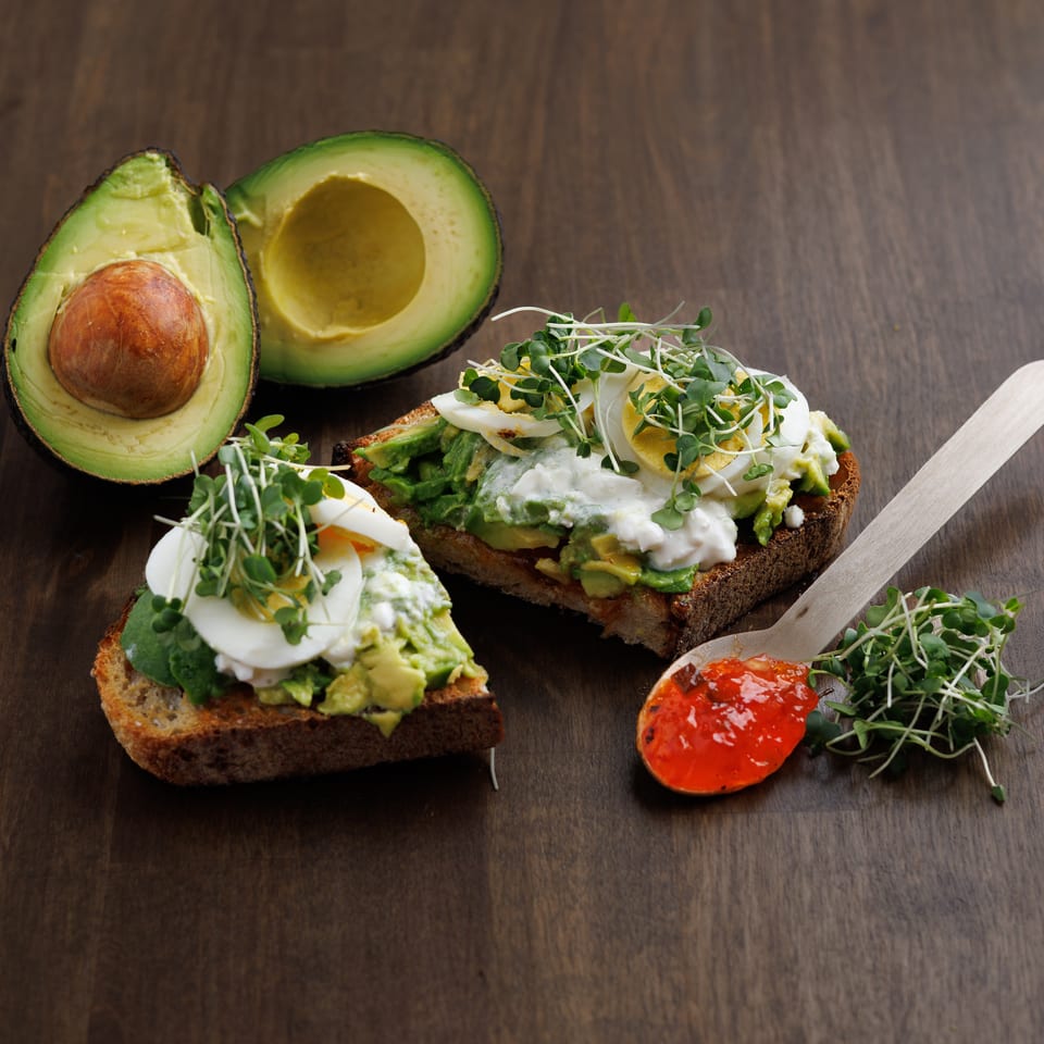 Avocado toast topped with poached eggs, creamy sauce, and microgreens beside halved avocados on a wooden table.