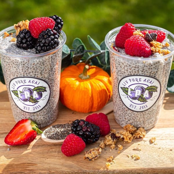 Two Liv Pure Acai chia puddings with berries and granola, a pumpkin, and loose fruit on a wooden surface.