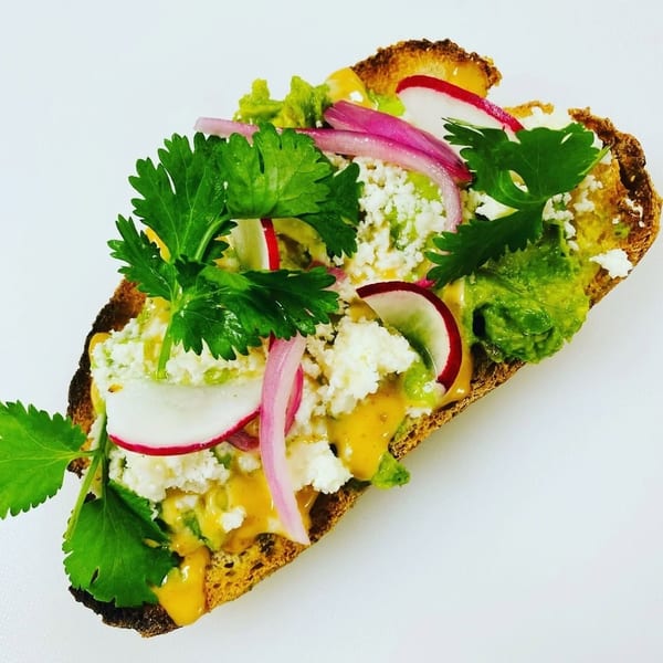 Avocado toast topped with crumbled cheese, red onion, radish slices, cilantro, and a drizzle of sauce.
