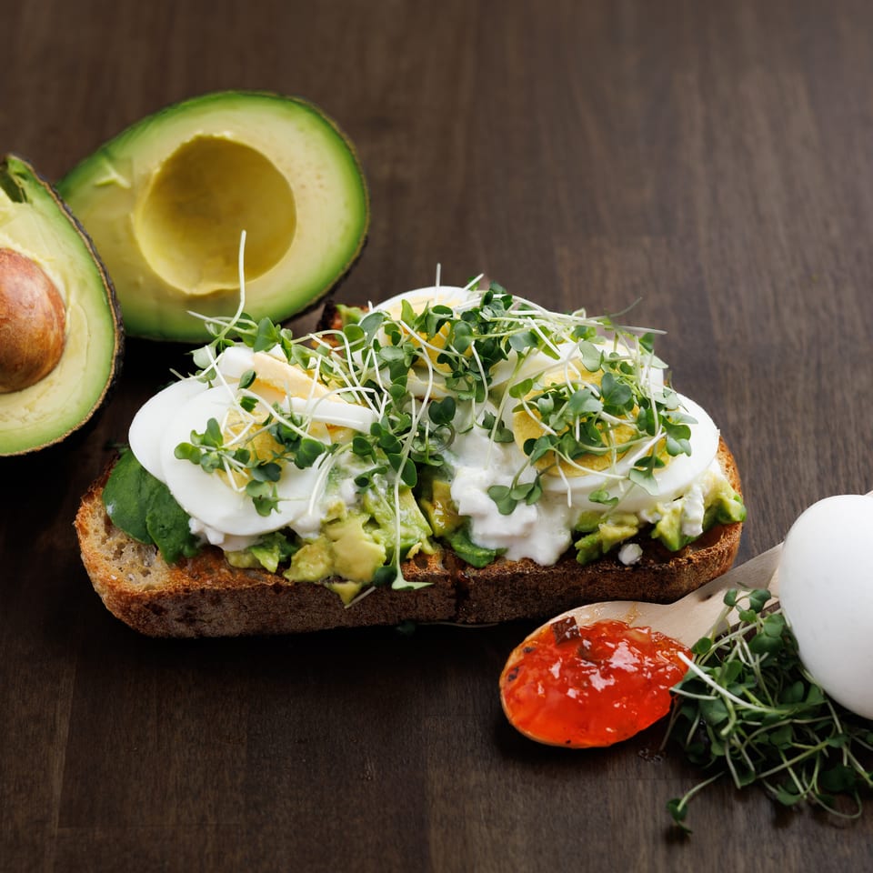Avocado toast with sliced eggs, microgreens, and feta cheese, with avocado halves and jam.