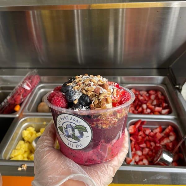 A hand in a clear glove holds a Liv Pure Acai bowl topped with berries, nuts, and granola.