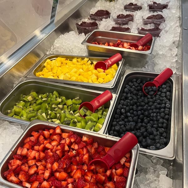 A vibrant fruit bar with containers of diced strawberries, kiwi, pineapple, blueberries, and acai bowls on ice.