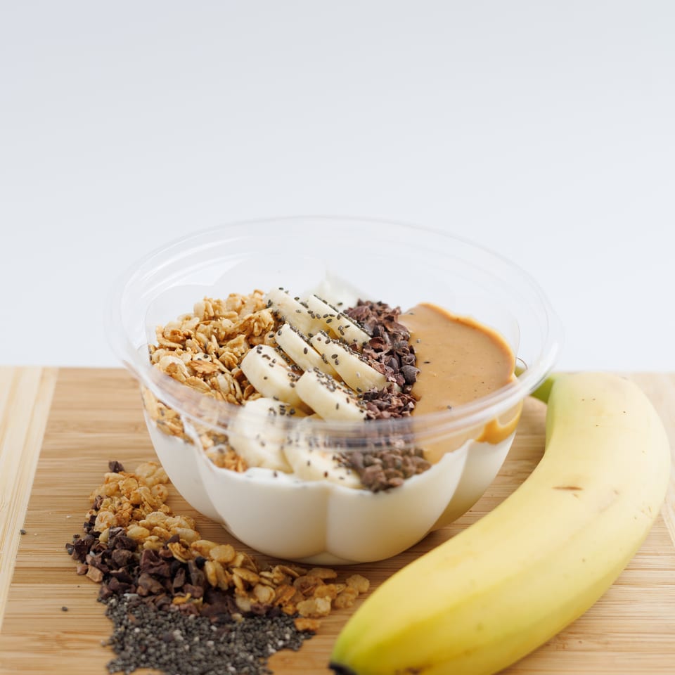 Bowl of yogurt topped with banana slices, granola, cacao nibs, chia seeds, and peanut butter on a wooden board.