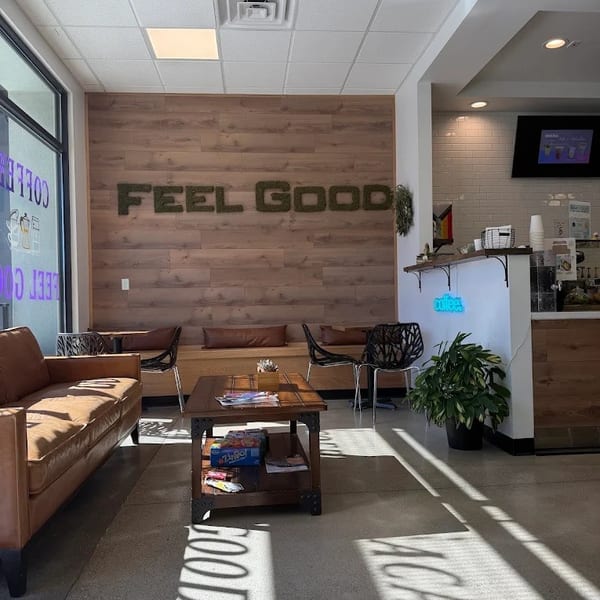 Cozy cafe interior with a "FEEL GOOD" moss sign, brown leather couch, and wooden coffee table.